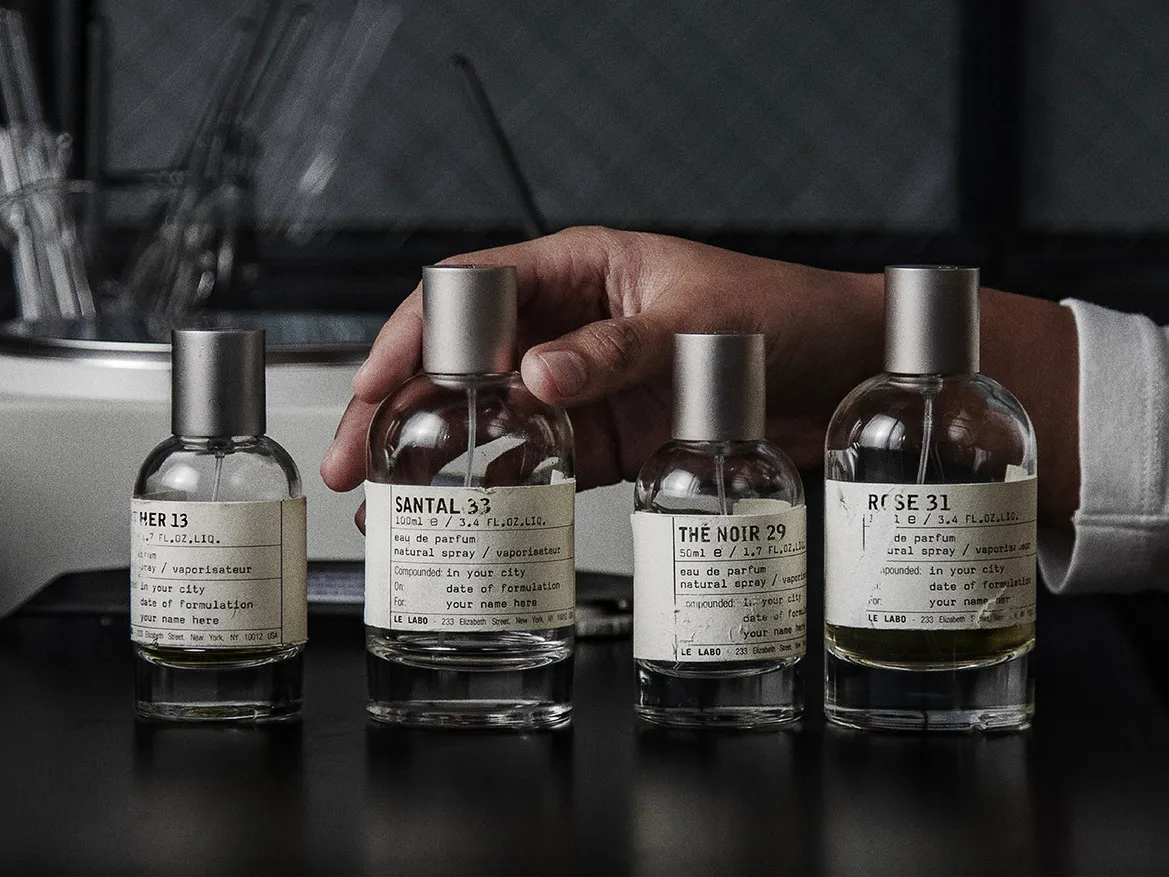 A hand arranging four Le Labo bottles on a dark counter